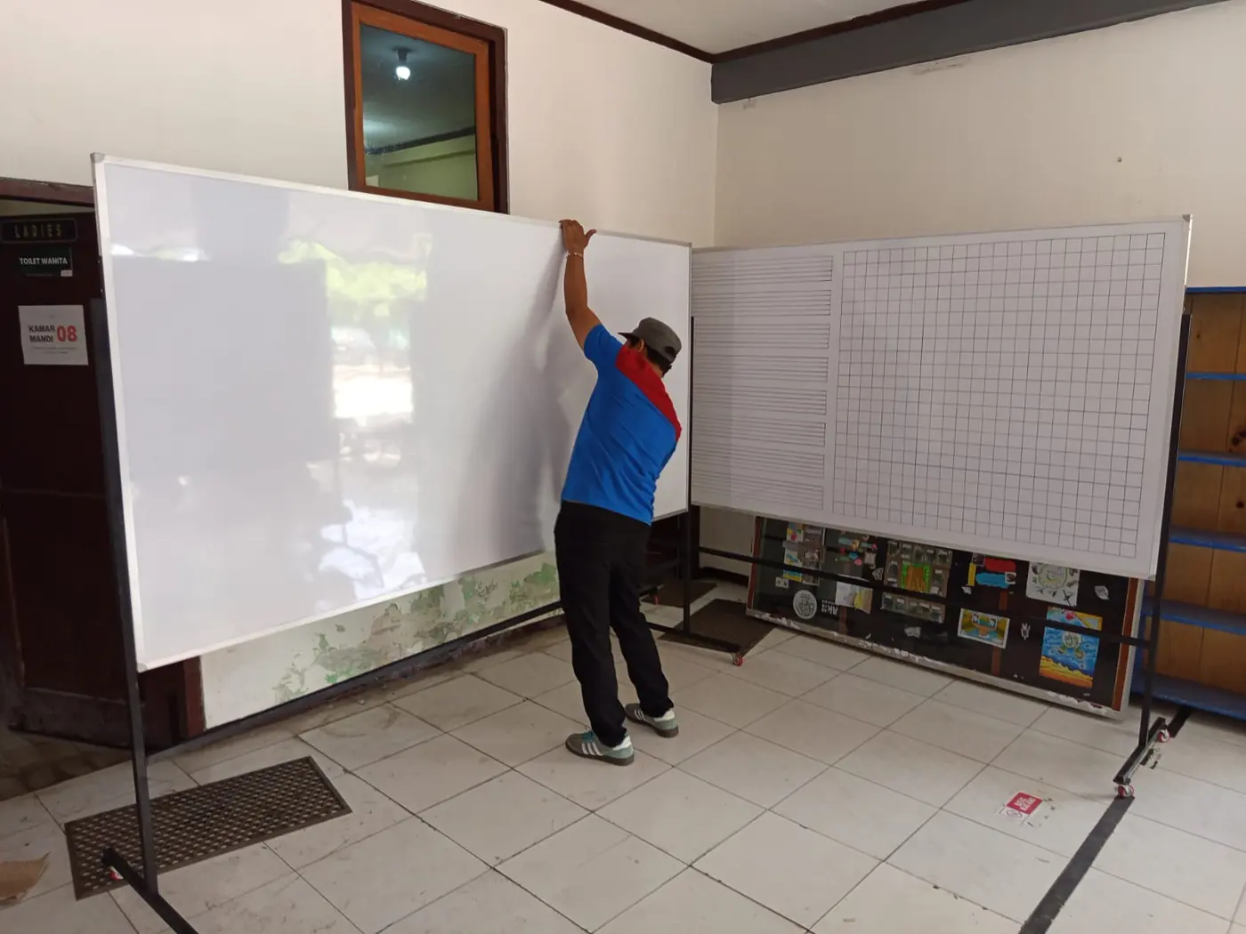 Pekerja merapikan papan tulis putih yang baru menempel di dinding ruang kelas