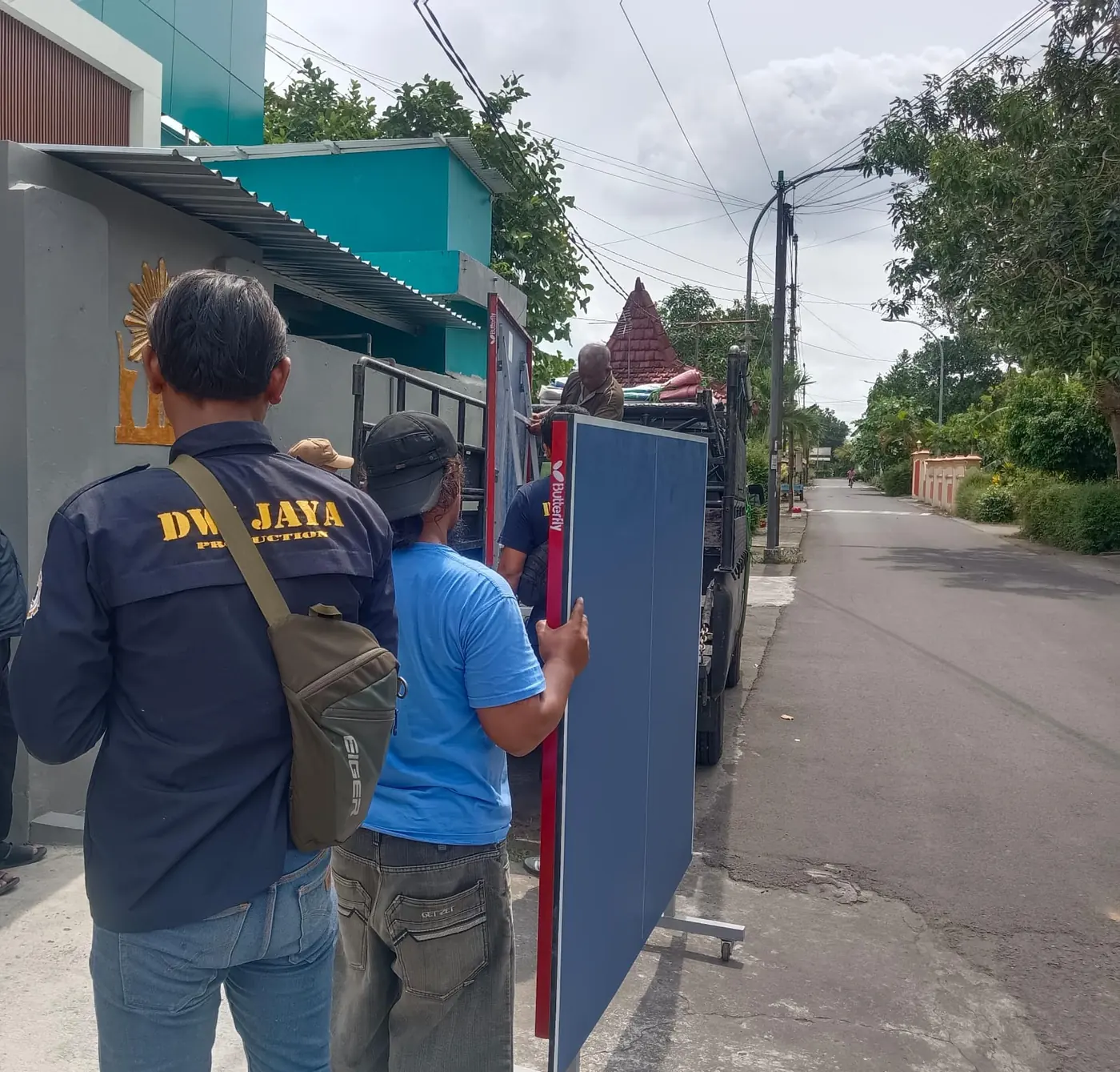 Dua staf Dwi Jaya membawa papan biru menyusuri jalan kampung menuju lokasi pemasangan